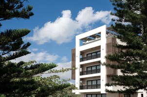Um edifício moderno de vários andares com varandas, rodeado por árvores e sob um céu limpo.
