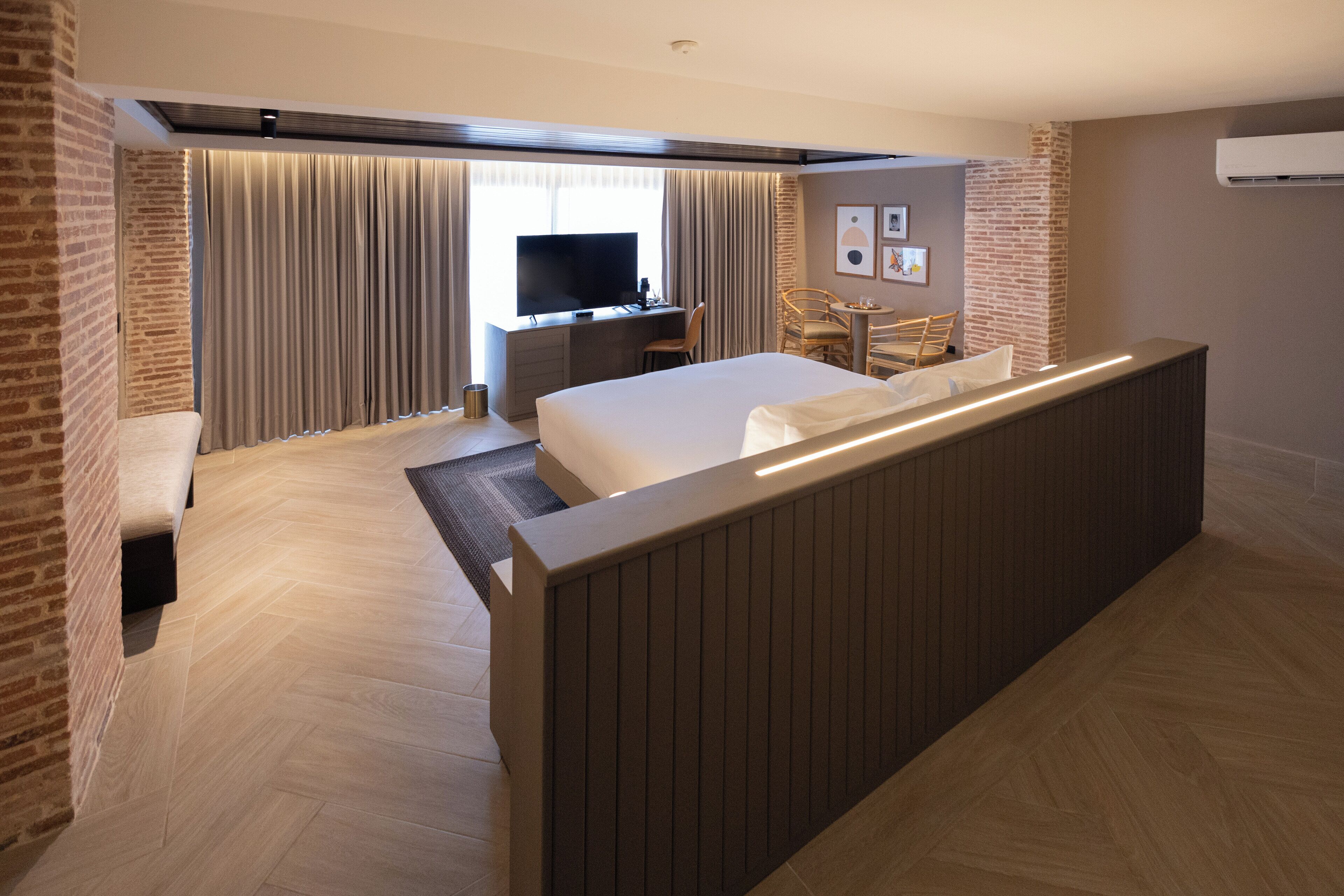 Quarto de hotel com cama grande, escrivaninha com TV, área de estar e parede decorativa de tijolos.