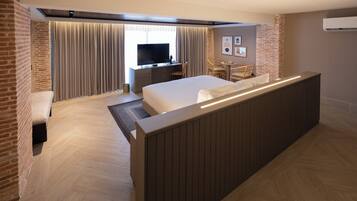 Quarto de hotel com cama grande, escrivaninha com TV, área de estar e parede decorativa de tijolos.