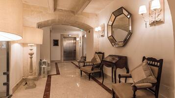 Un hall d’hôtel avec un sol en marbre, un comptoir de réception, un espace d’attente avec des fauteuils, un grand miroir et un escalier menant à un ascenseur.