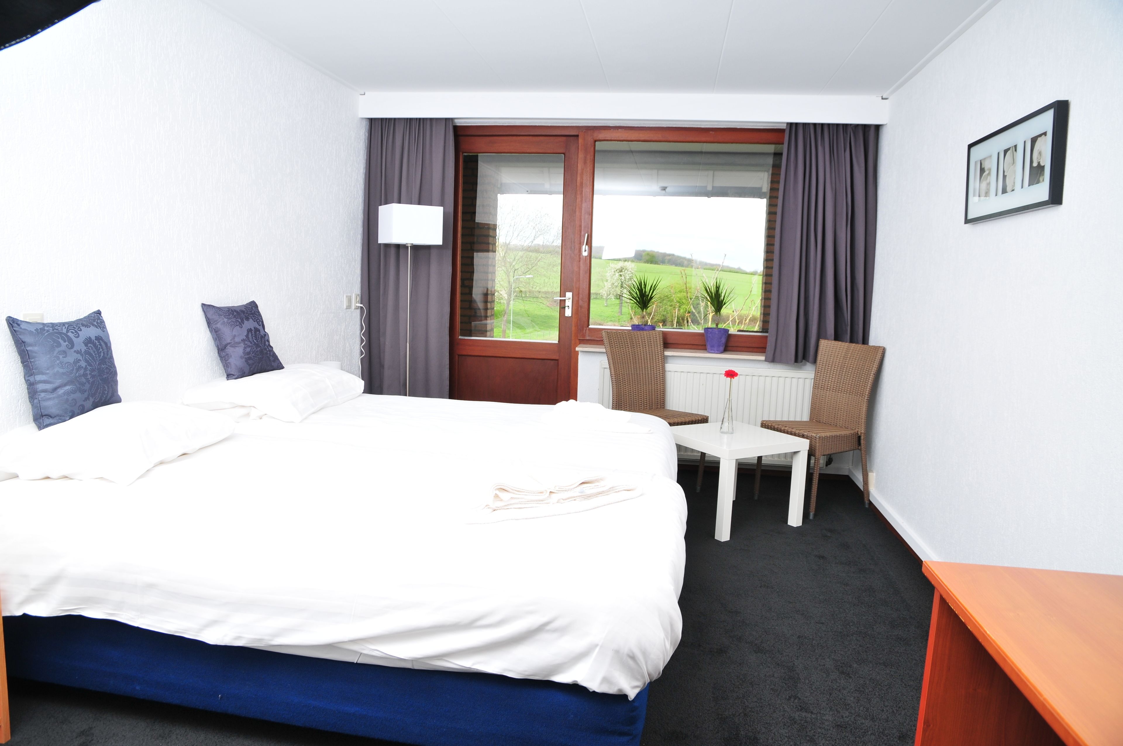 Quarto de hotel com uma cama, uma pequena mesa, uma cadeira, uma janela com cortinas e vista para o verde lá fora.