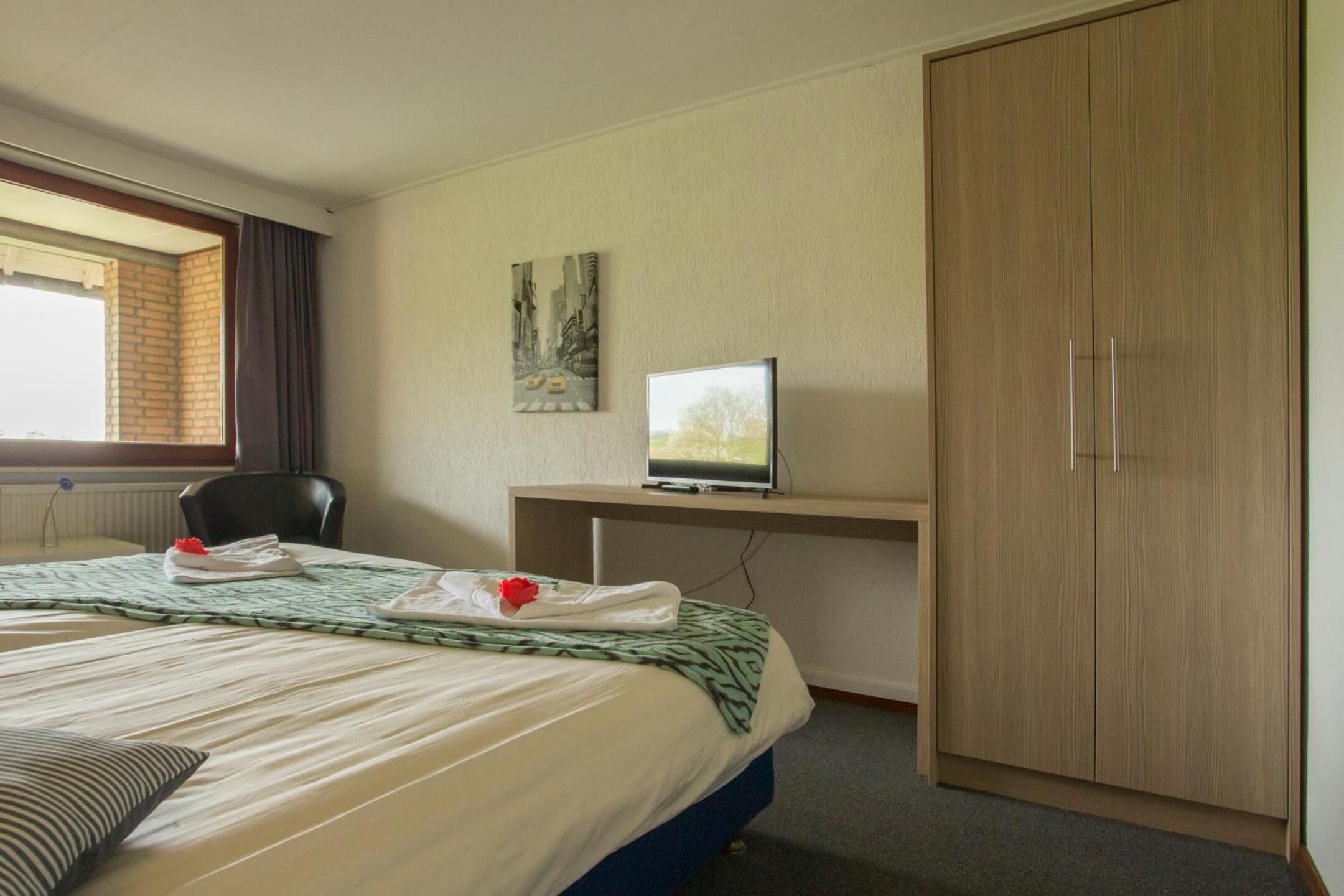 Quarto de hotel com cama, escrivaninha com televisão, guarda-roupa e janela com cortinas.