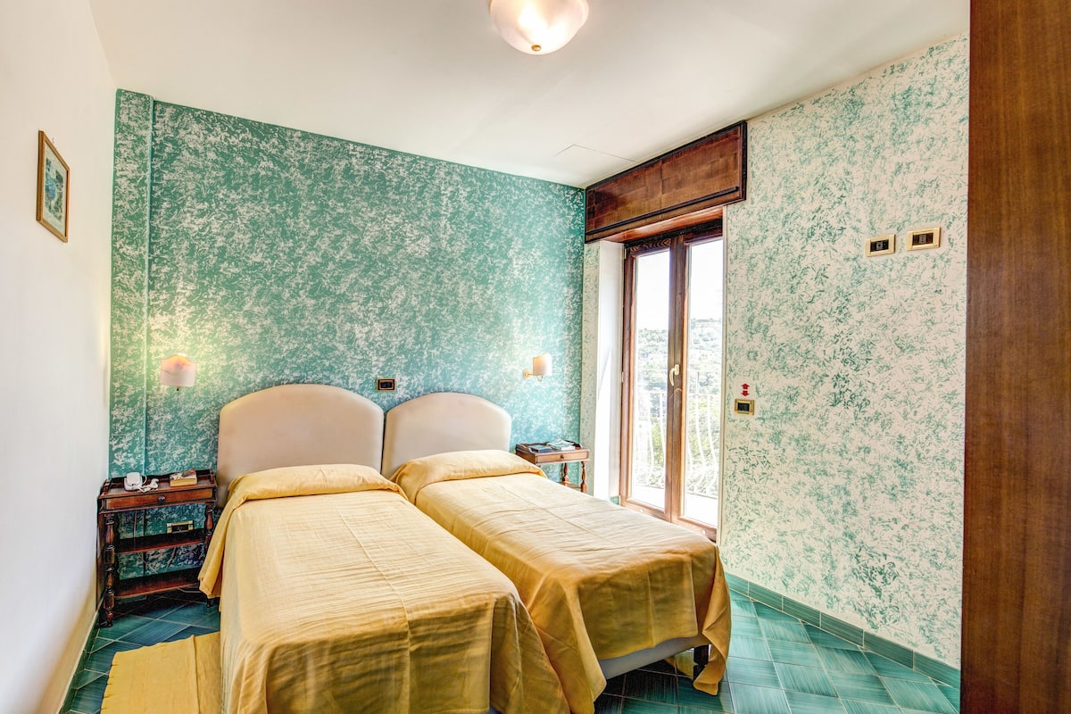 Ein Zimmer mit zwei Betten, einem hölzernen Nachttisch, einem Fenster mit Blick und einer Wand mit Blumenmuster.
