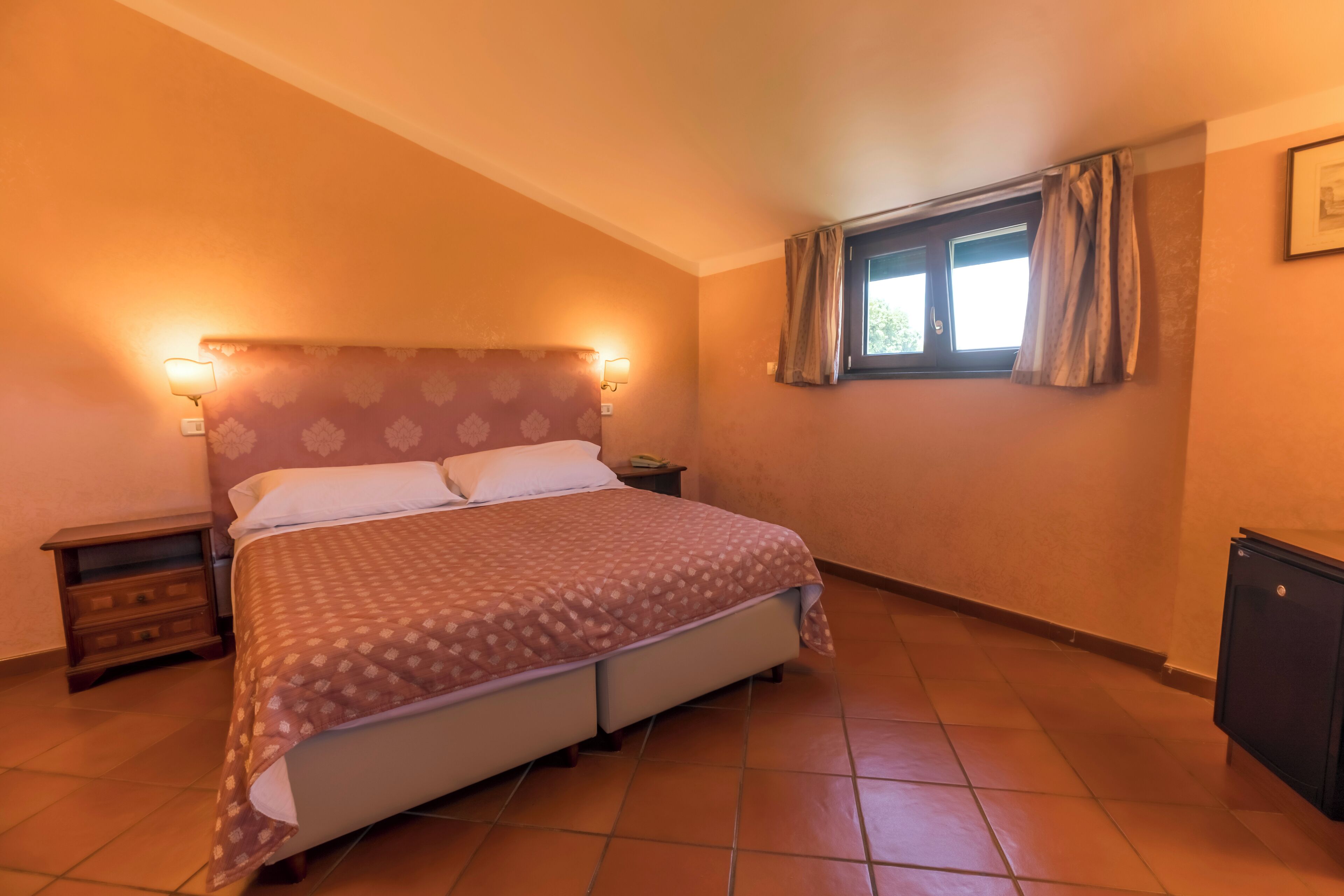 Quarto com cama, mesas de cabeceira, janela com cortinas e radiador.
