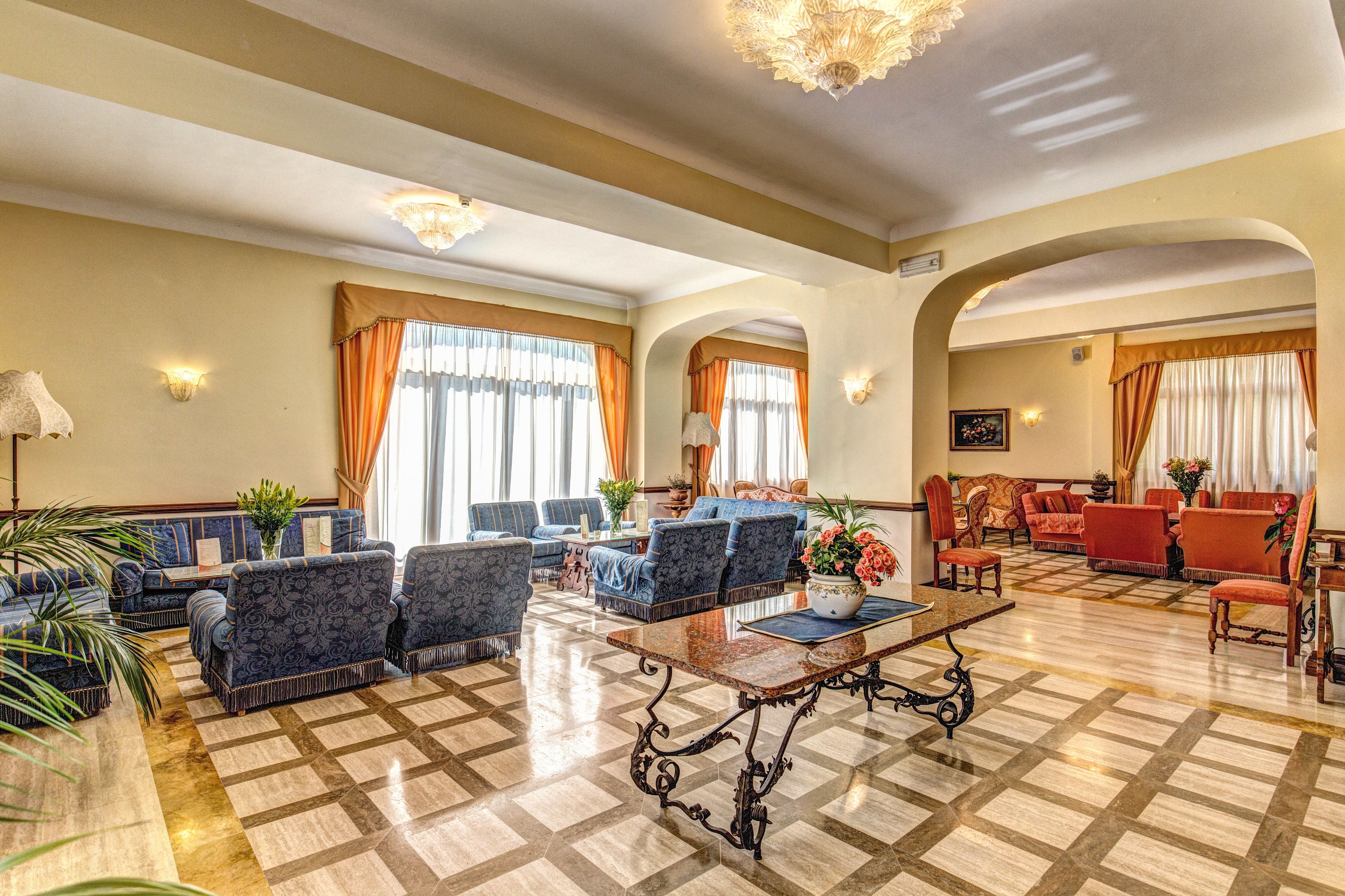Um hall amplo com piso xadrez, sofás azuis estampados, uma mesa de centro e um lustre.