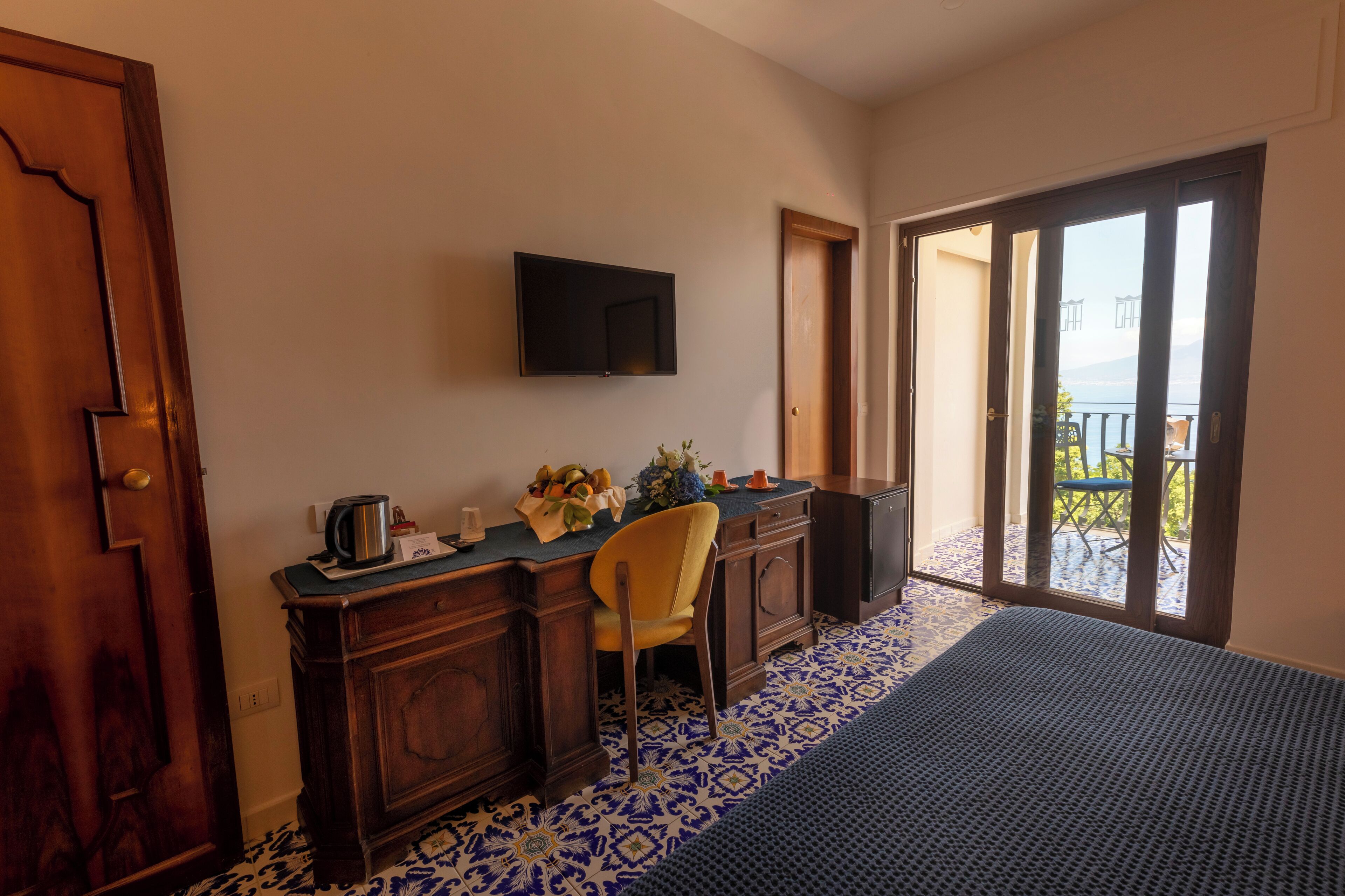 A hotel room with a wooden desk, a television mounted on the wall, a chair, and a balcony with a view.