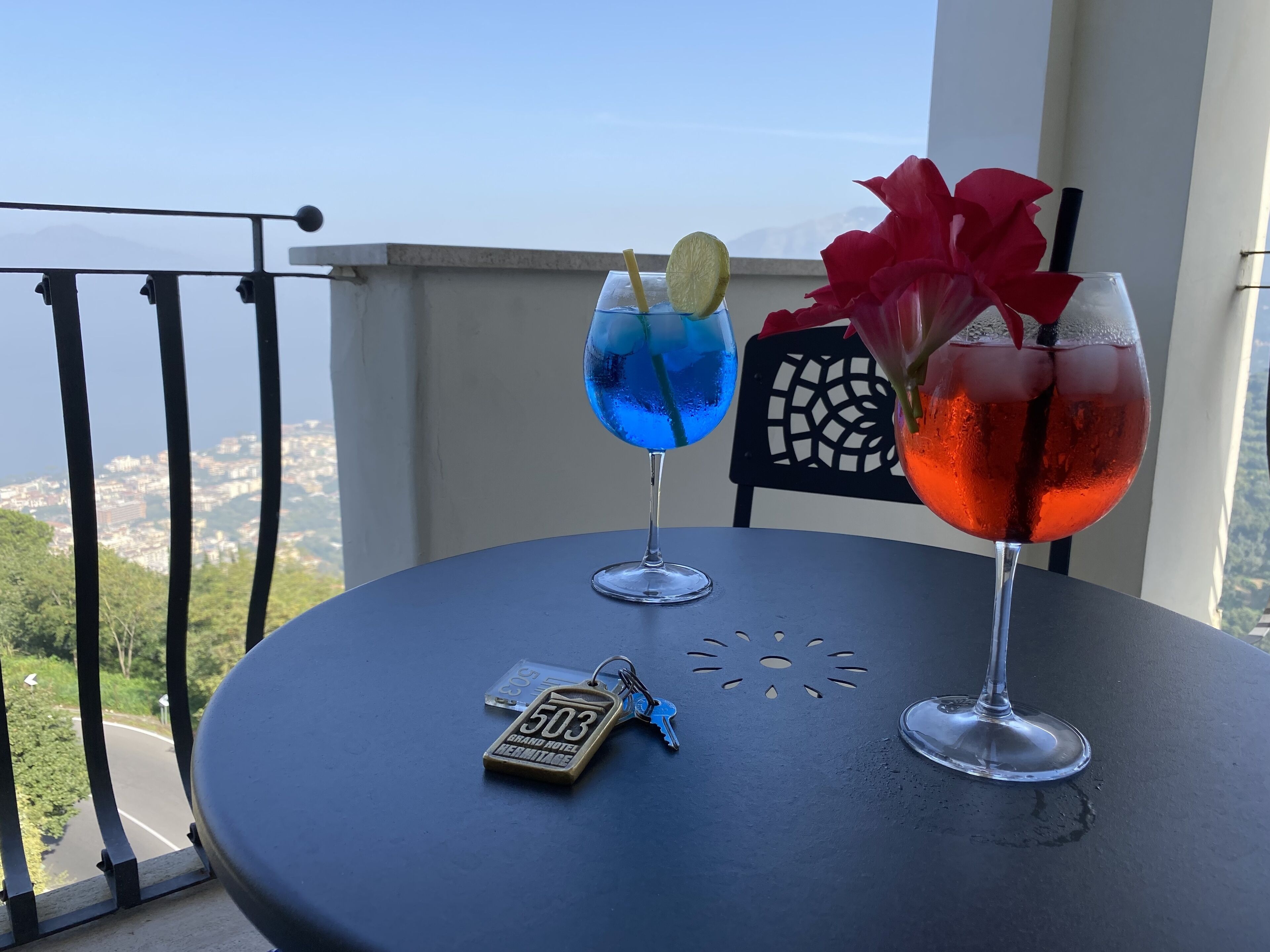 A table with two drinks and a keychain on a balcony overlooking a cityscape.