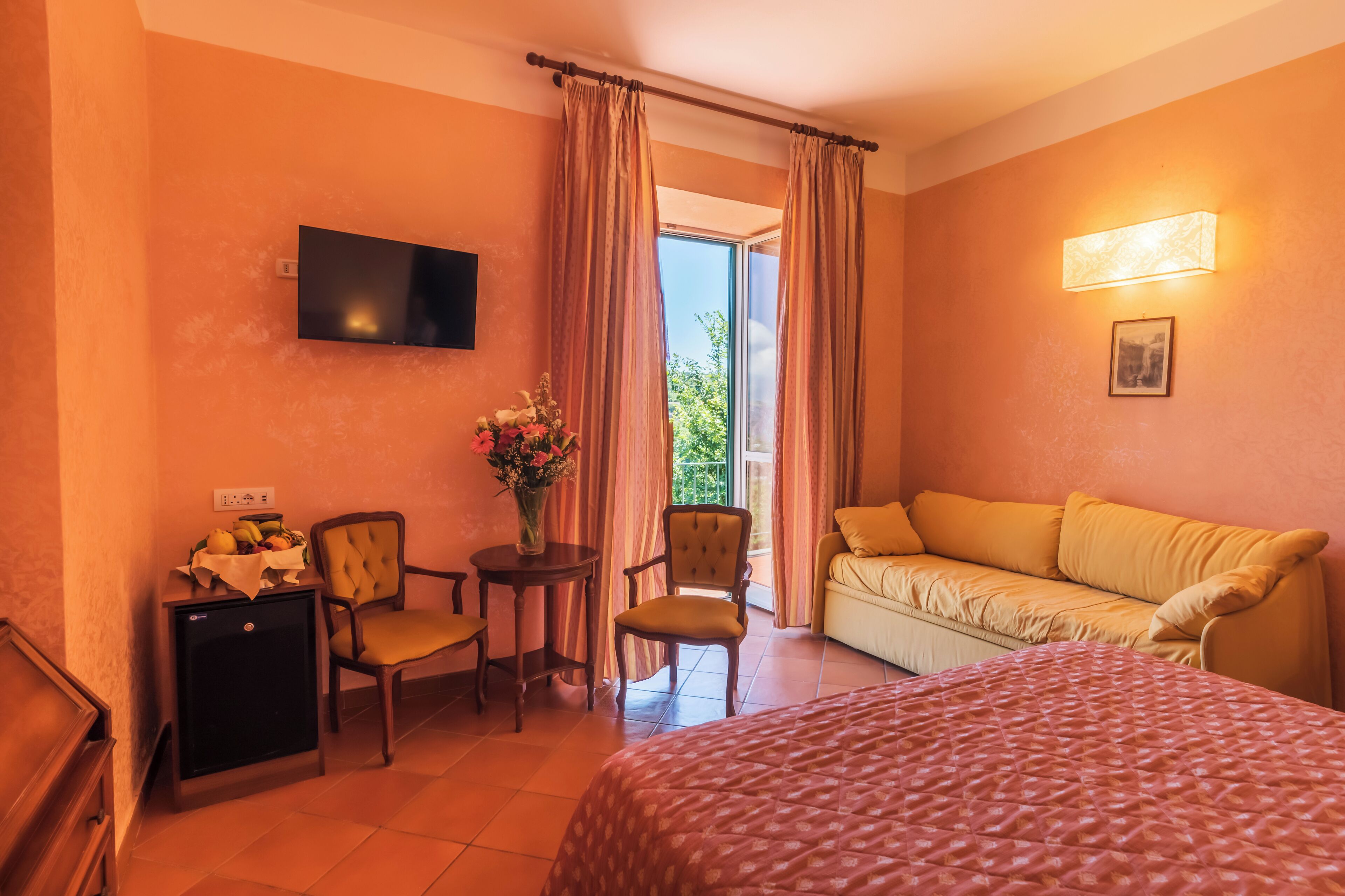 Habitación de hotel con cama, sofá, televisión, una mesita con un jarrón de flores y una silla.