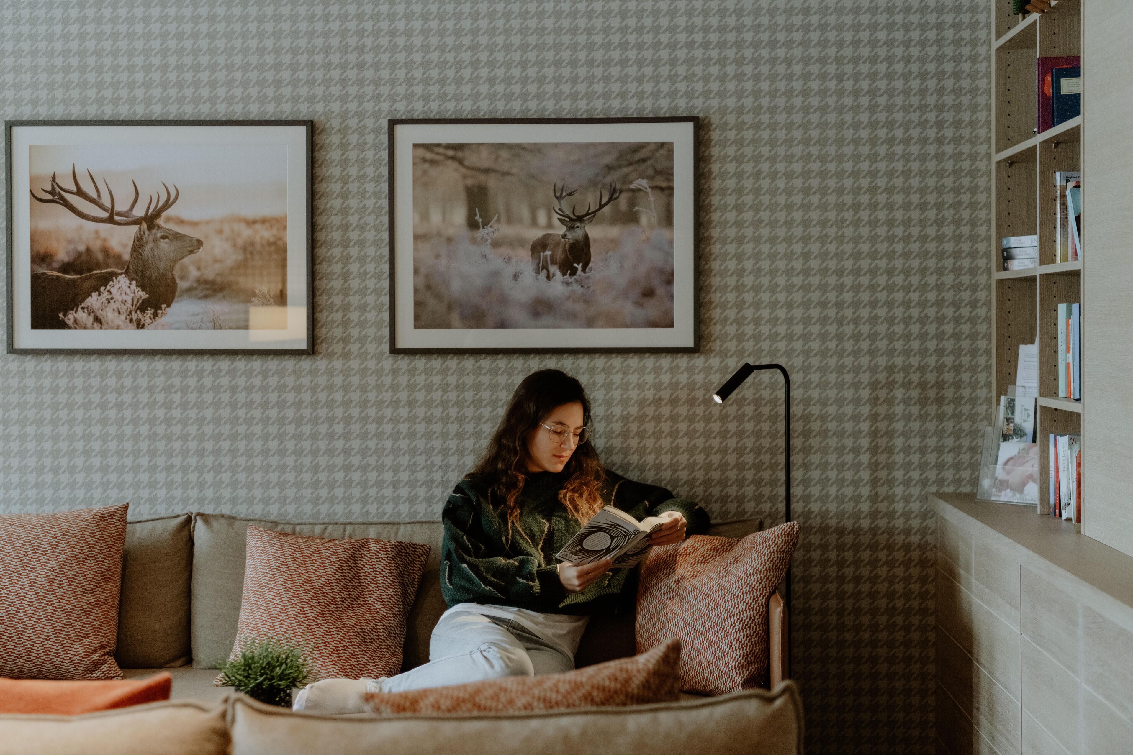 A person sitting on a couch reading a book, with two framed deer pictures on the wall behind them.