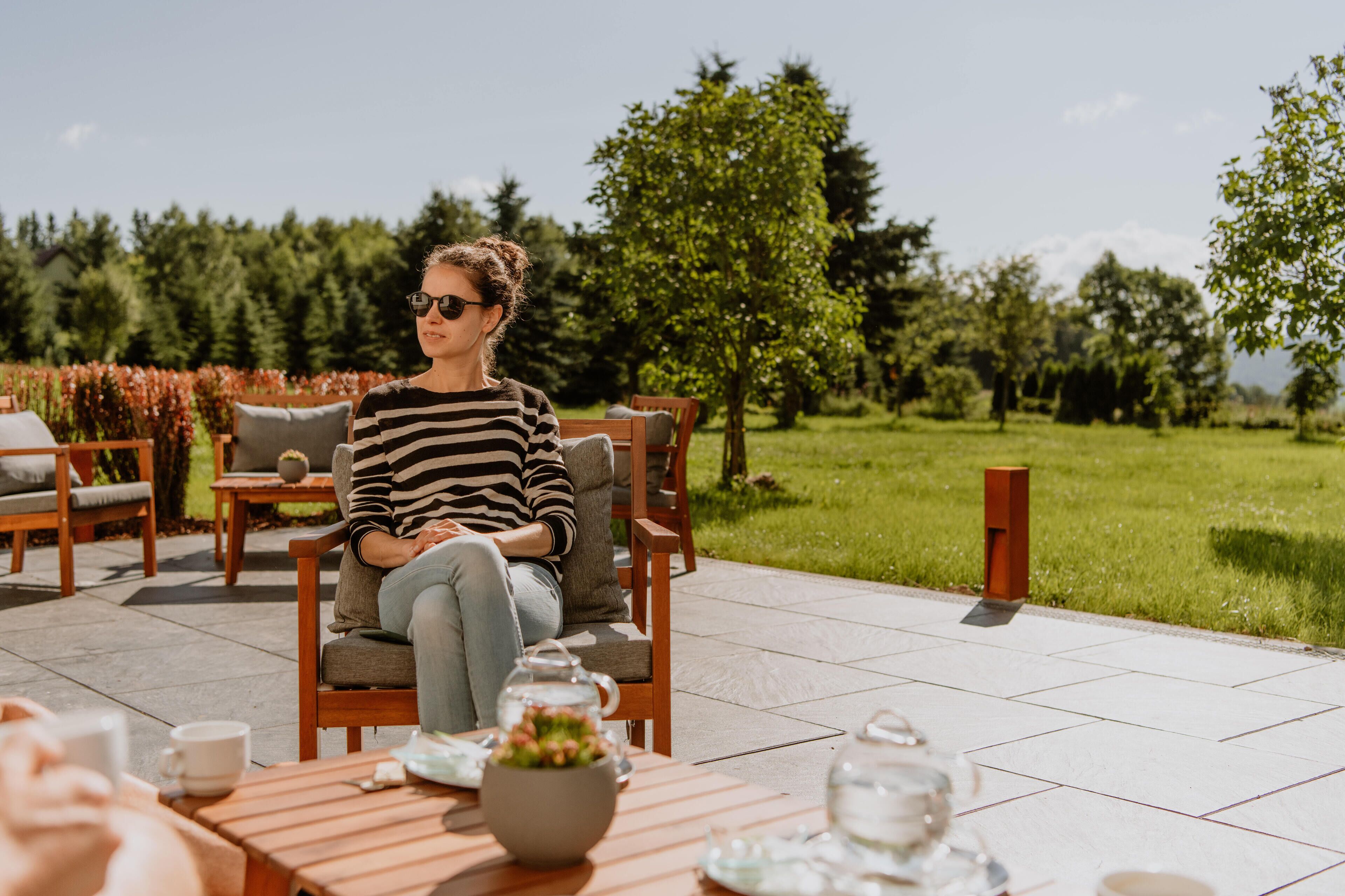 A person seated on a wooden chair outdoors with a table in front, surrounded by greenery and trees.