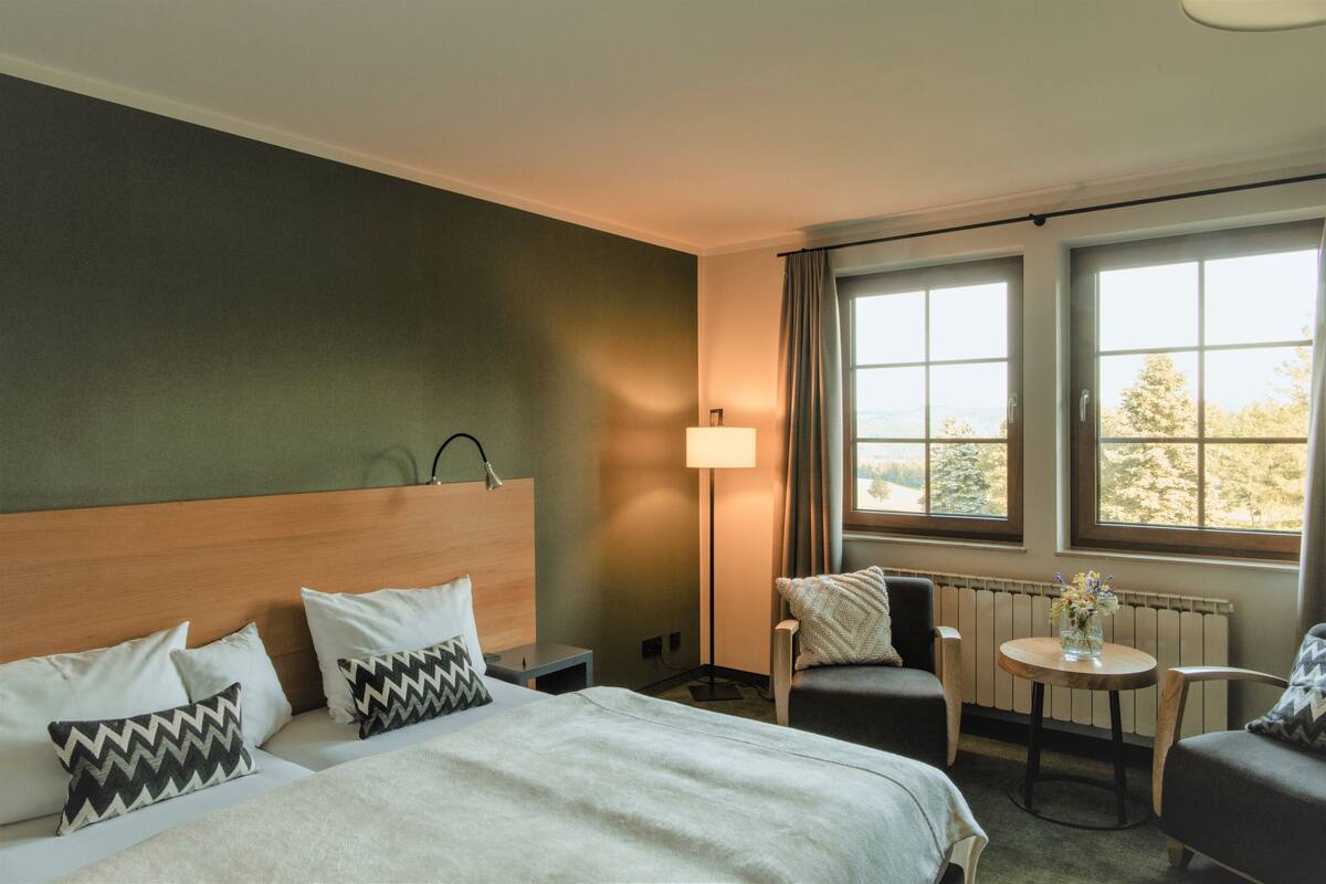 Een hotelkamer met een bed, twee stoelen, een kleine tafel met een vaas en een raam met gordijnen.