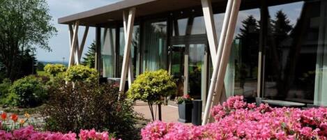 A modern building with a glass facade and a flat roof, surrounded by well-maintained gardens and shrubs.