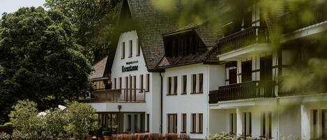 A traditional building with a sign reading "Kreuztanne." It features a balcony, multiple windows, and a well-maintained garden with trees and shrubs.