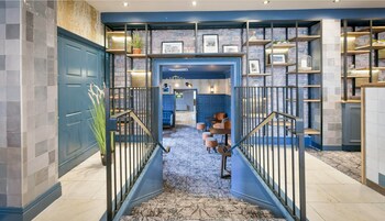 Un interior moderno con una puerta azul que da a un pasillo con piso estampado, una escalera con barandillas metálicas y una pared de ladrillo con estantes que exhiben diversos objetos.