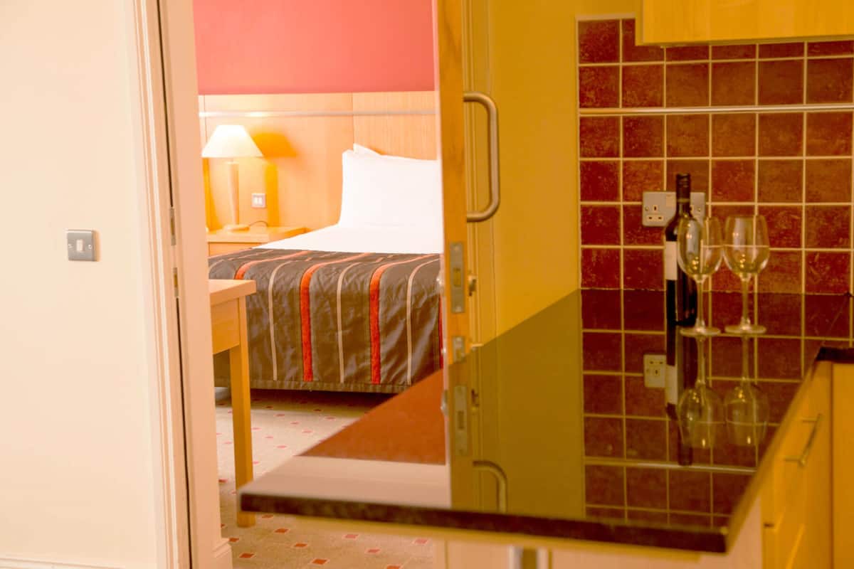 Ein Hotelzimmer mit Bett, Nachttisch, Lampe und einer Küchenzeile mit Weingläsern.