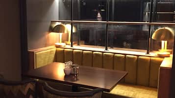 A cozy dining area with a wooden table and two upholstered chairs, a cushioned bench, and a large window with metal frames.
