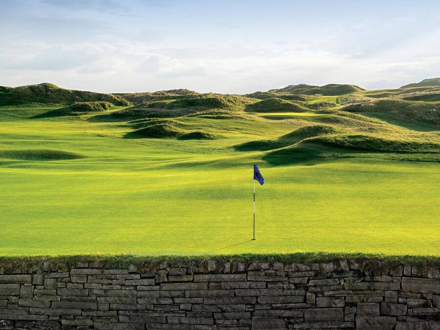 A golf course with rolling green hills and a stone wall at the foreground.