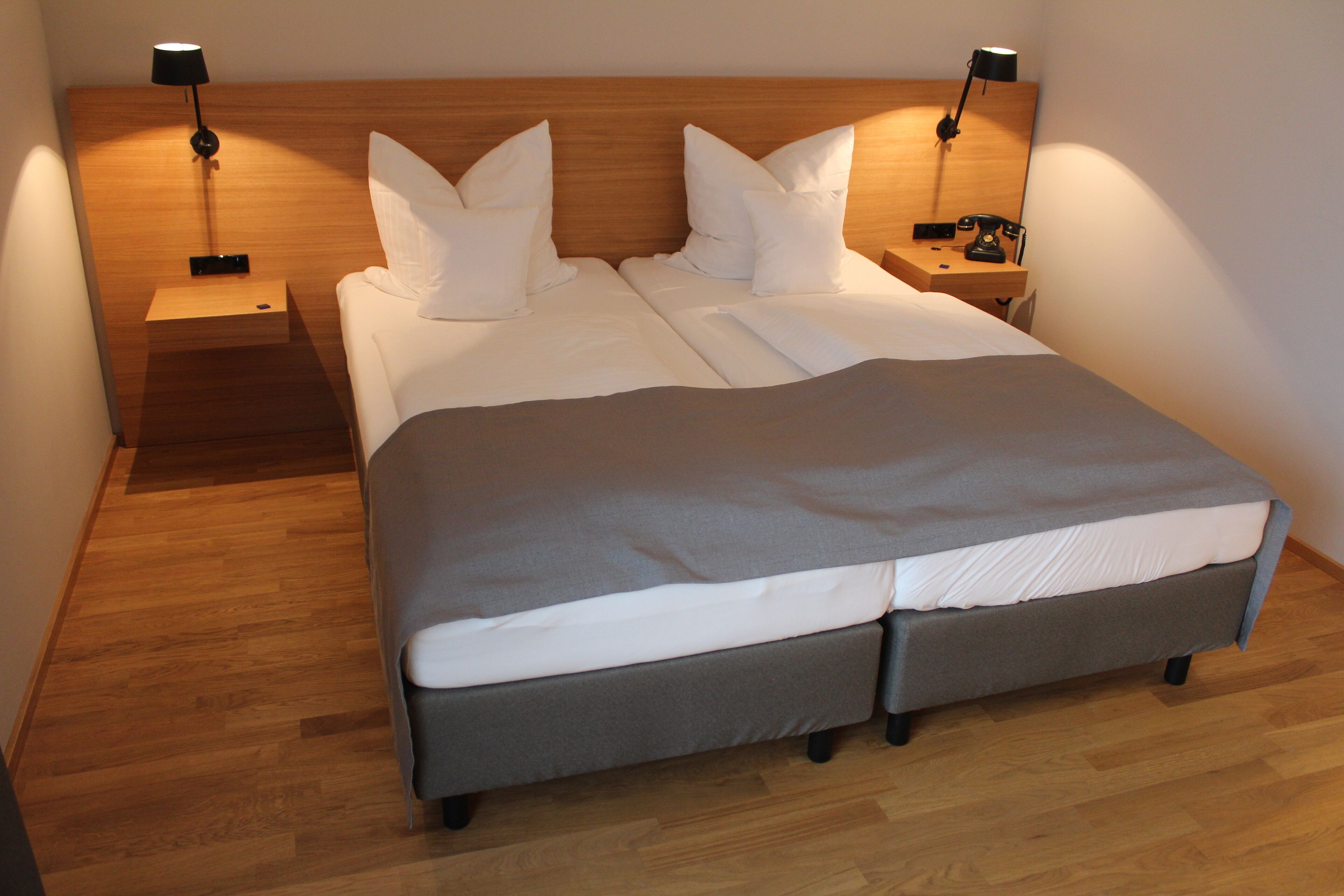 Cama doble con colcha gris y ropa de cama blanca, dos mesitas de noche con lámparas y un cabecero de madera.