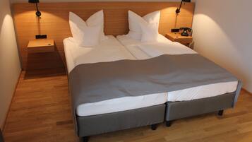 Cama doble con colcha gris y ropa de cama blanca, dos mesitas de noche con lámparas y un cabecero de madera.