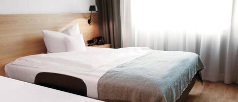 Habitación de hotel con cama, mesita de noche, lámpara y vistas al exterior a través de cortinas.