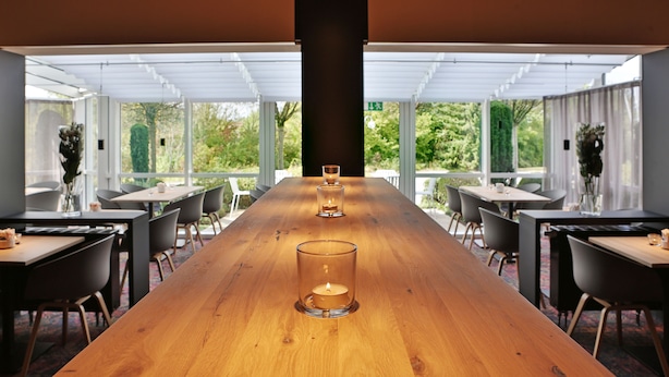 A long wooden table with candles in a modern dining area with large windows and chairs. - Hotel Stadt Balingen (Balingen)