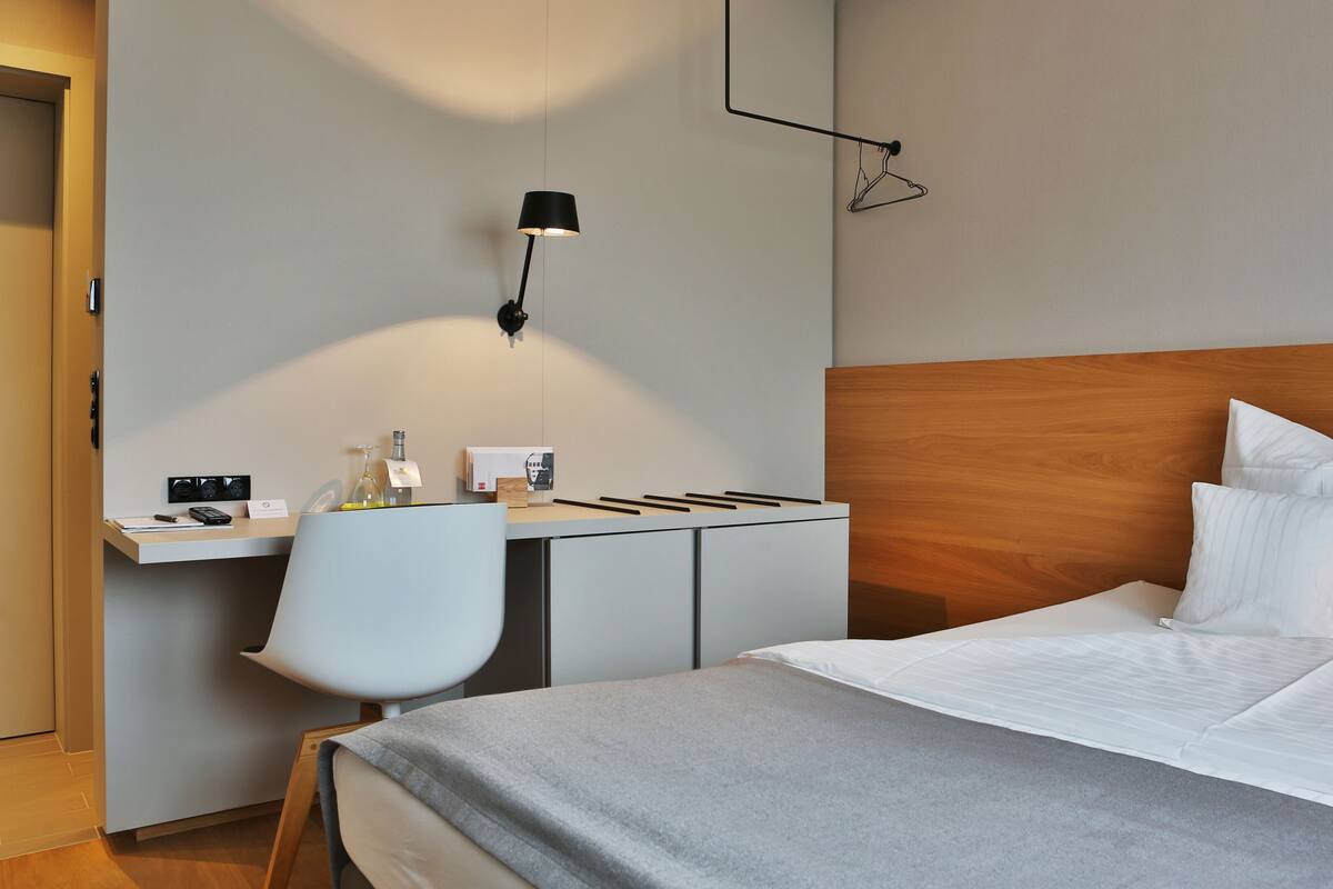 A hotel room with a bed, a desk, a chair, and a wall-mounted lamp.