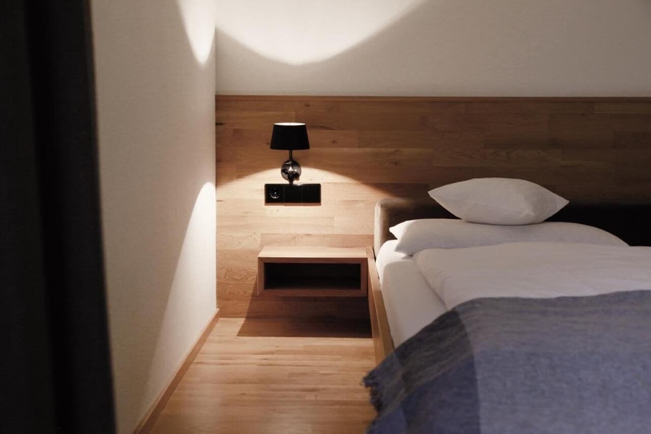 A bedroom with a wooden headboard, a bedside table with a lamp, a bed with white bedding, and a small wooden shelf.