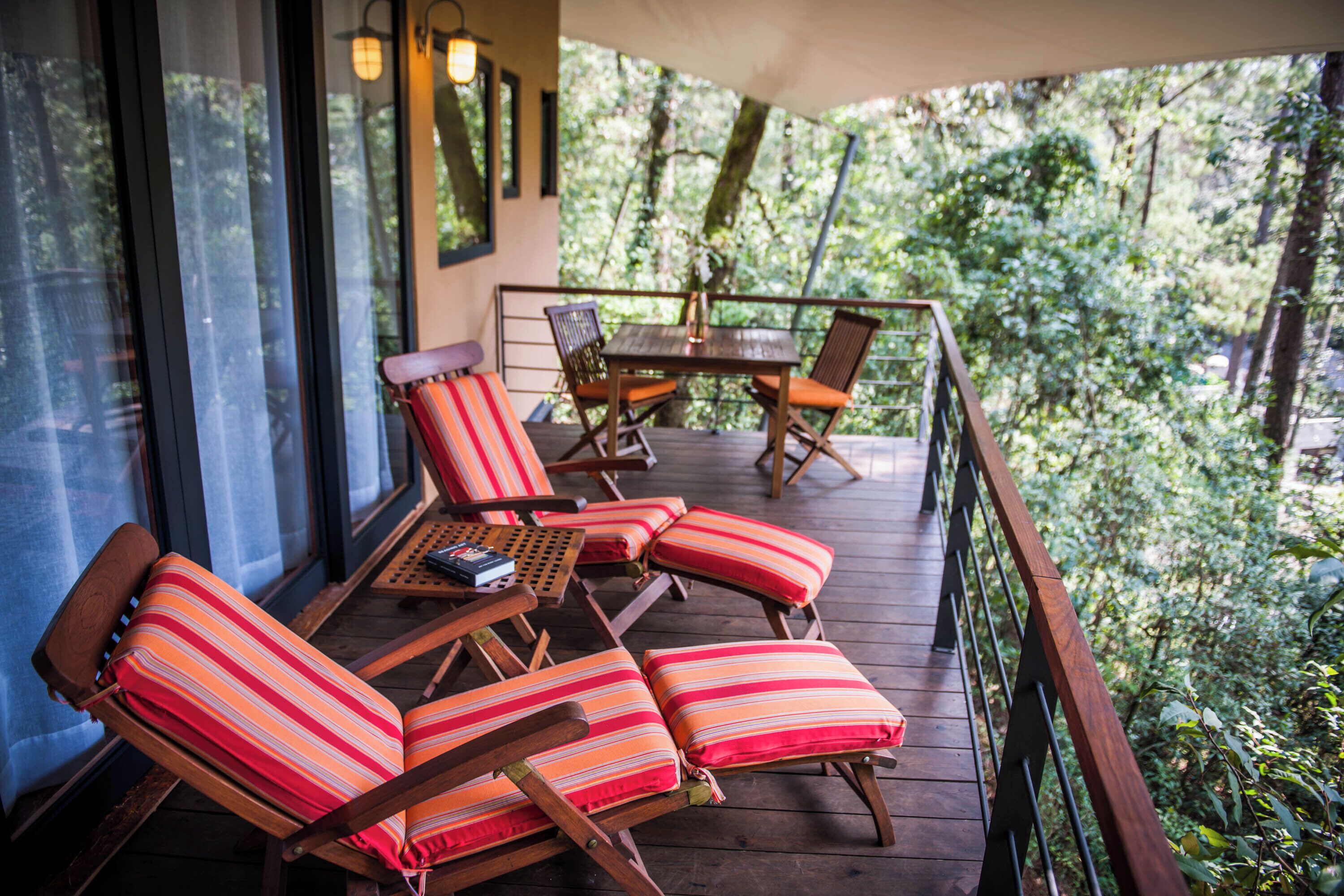 Terraza de madera con sillones de descanso y una mesa, rodeada por una barandilla de madera, con vista a un área boscosa.
