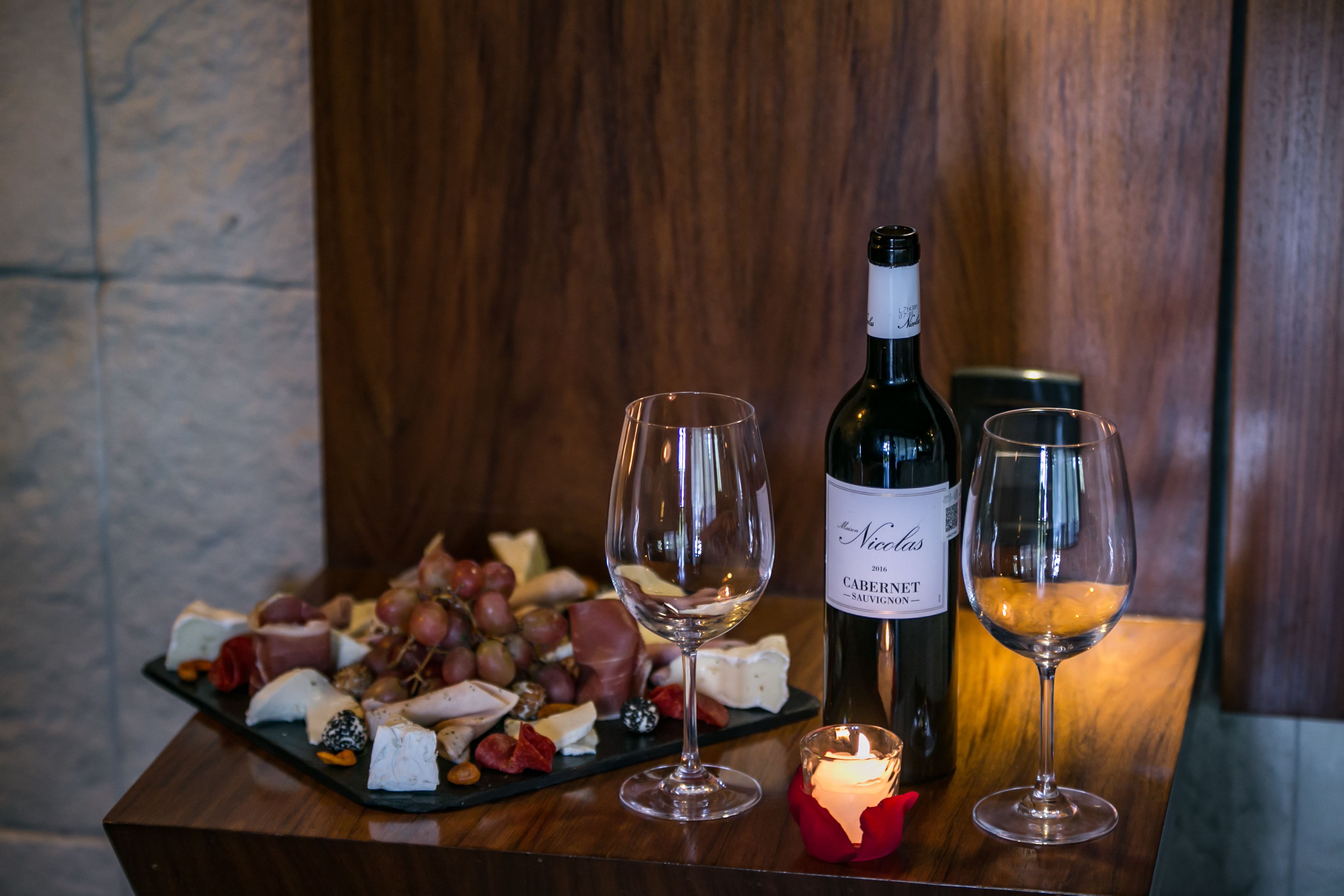 A wooden table with a bottle of wine, two glasses, and a platter of cheeses and grapes.