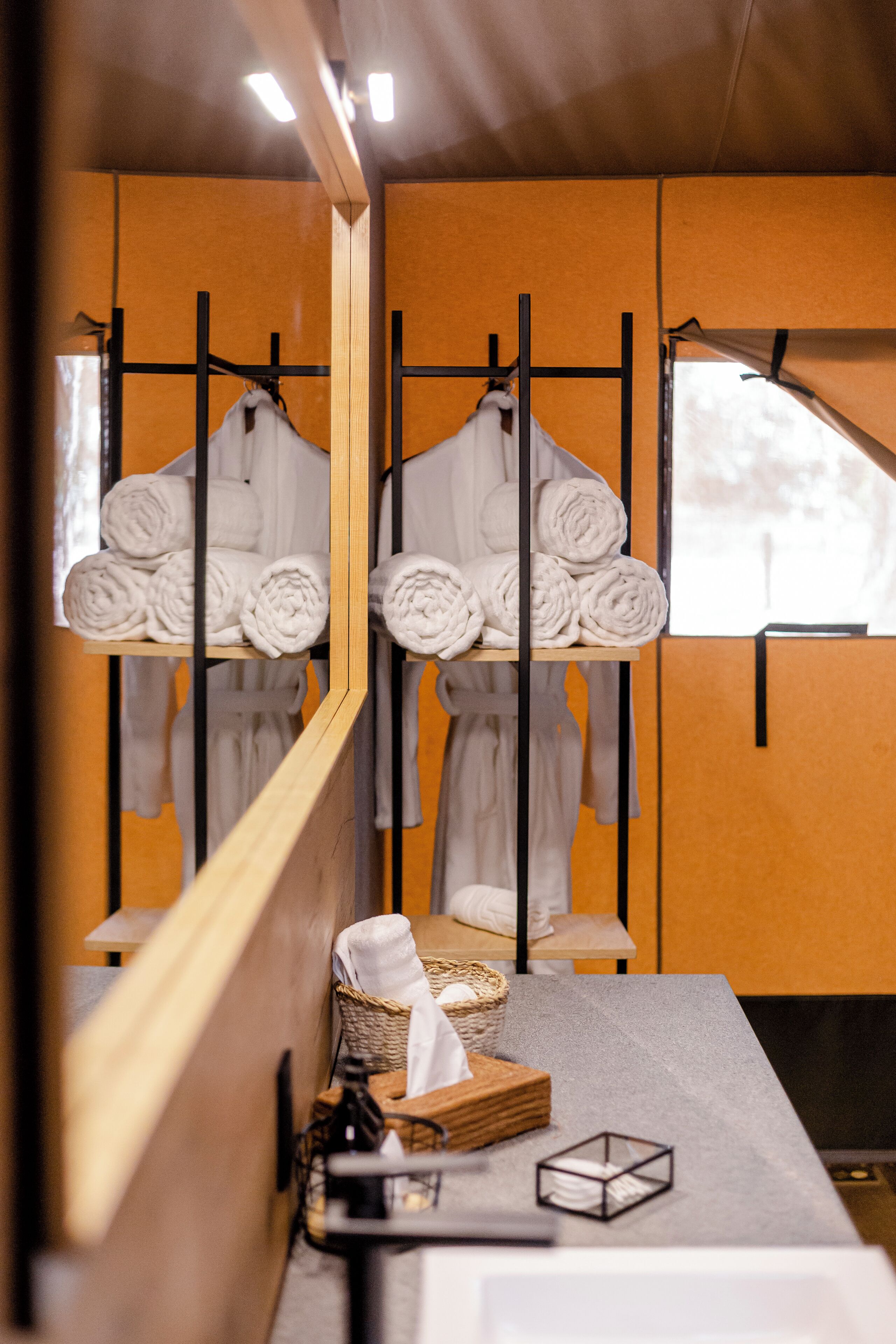 Un baño con un banco de madera, un perchero con toallas enrolladas, una cesta y una mesita con una vela y pañuelos.