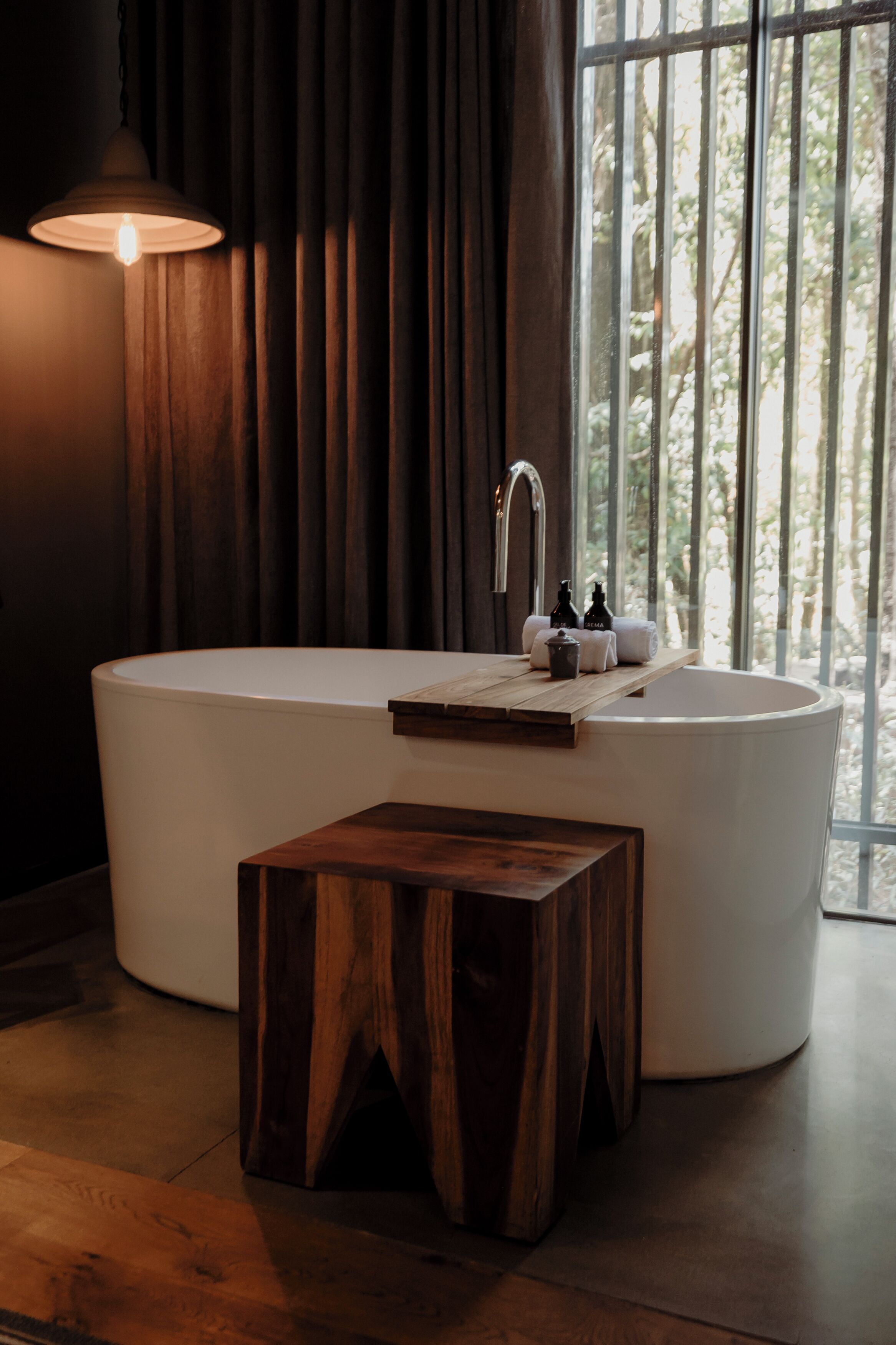 Un baño moderno con una bañera independiente, una mesita de madera y un ventanal con vistas a árboles.