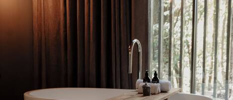 Un baño moderno con una bañera independiente, una mesita de madera y un ventanal con vistas a árboles.