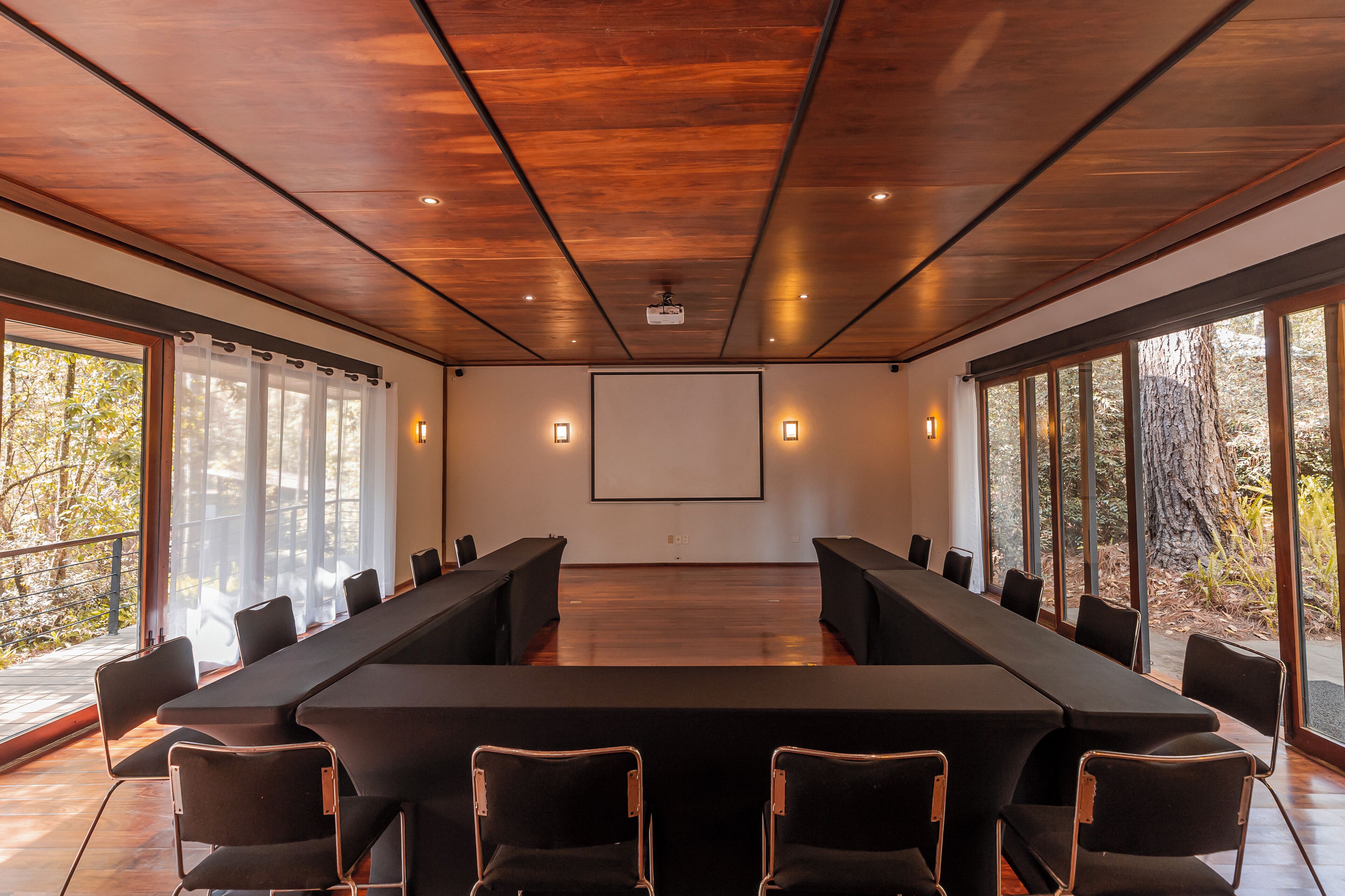 A conference room with a long table, chairs, a projector screen, and large windows with a view of trees.
