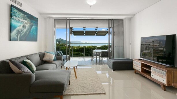 A modern living room with a grey sofa, a flat-screen TV, and a view of the outdoors. - Cairns Private Apartments (Cairns)