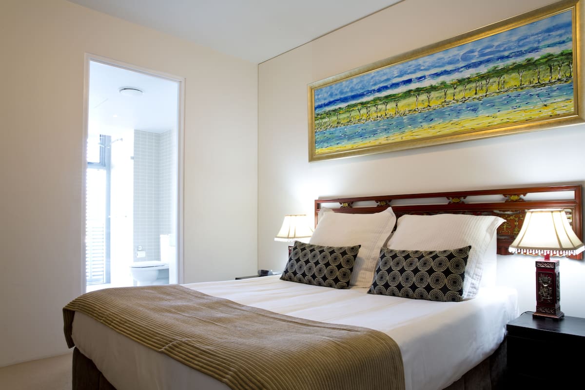 A hotel room with a large bed, two bedside lamps, a painting above the bed, and a view of the bathroom through an open door.