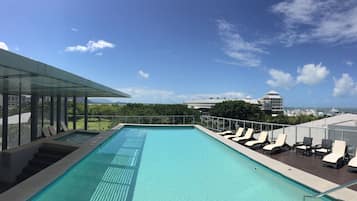 A rooftop infinity pool with lounge chairs and a glass balustrade overlooking a marina and cityscape.