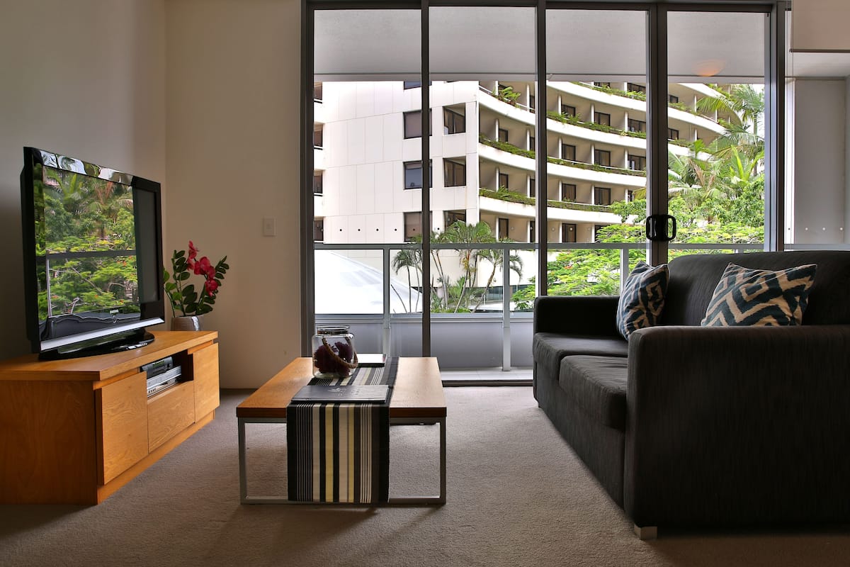 A modern living room with a television, a coffee table, a sofa, and a large window offering a view of a multi-story building and greenery.