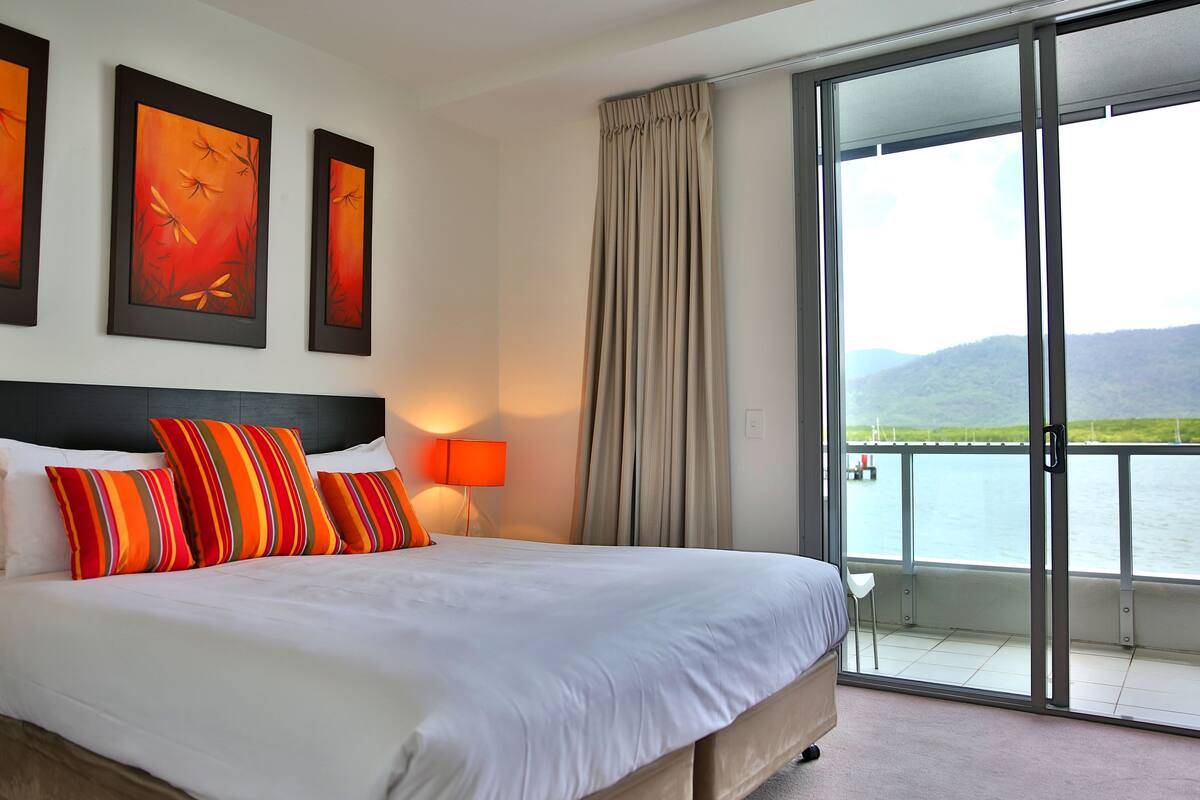 A hotel room with a large bed, a view of a body of water, and artwork on the wall.