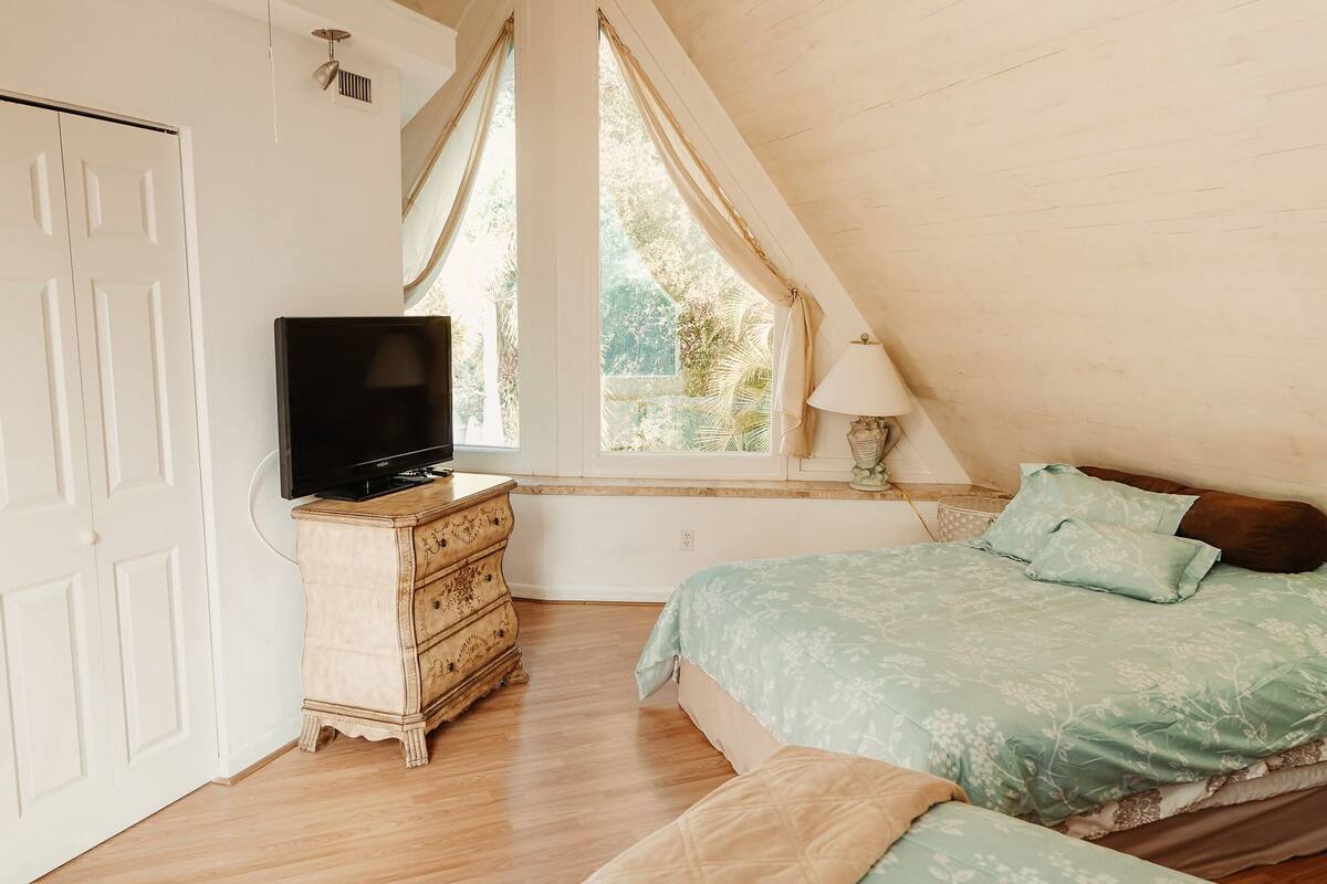 A bedroom with a wooden dresser, a television, a bed with a green floral bedspread, and a window with curtains.