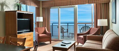 A hotel room with a balcony, a TV, a dining table, and two sofas.