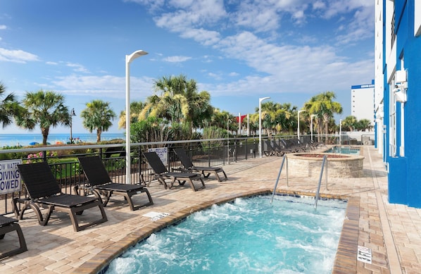 A pool area with lounge chairs, a hot tub, and a view of the ocean.