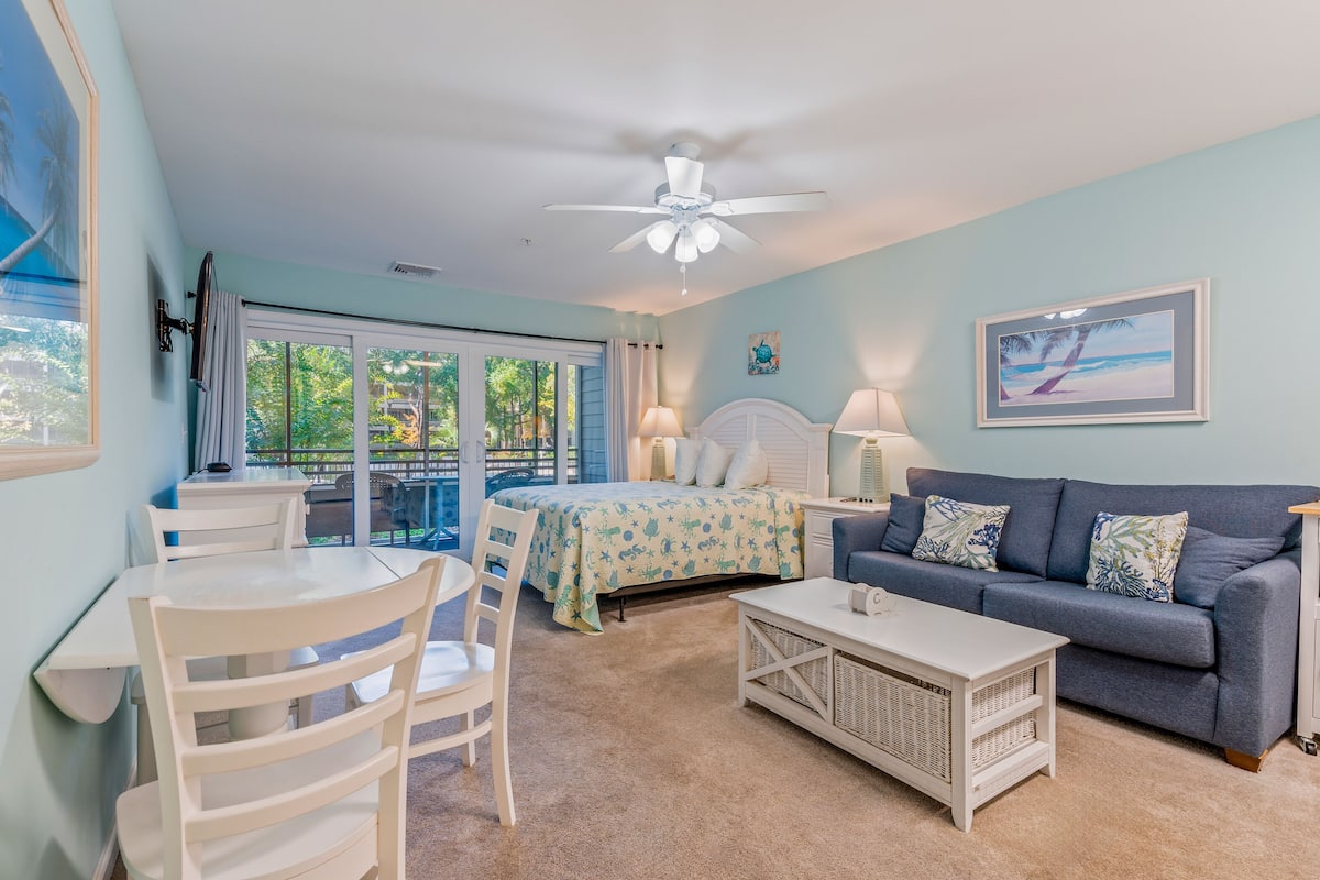 A bedroom with a bed, a blue sofa, a white coffee table, and a dining area with chairs and a table.