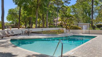 A rectangular swimming pool with a tiled edge, surrounded by a paved deck with lounge chairs and tables.