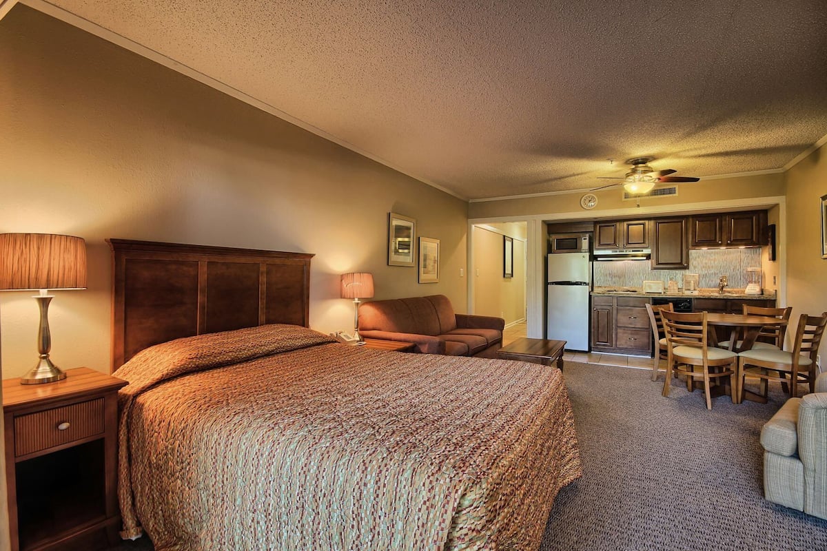 A bedroom with a large bed, bedside lamps, a nightstand, a wooden headboard, a sofa, and a dining area with a table and chairs.
