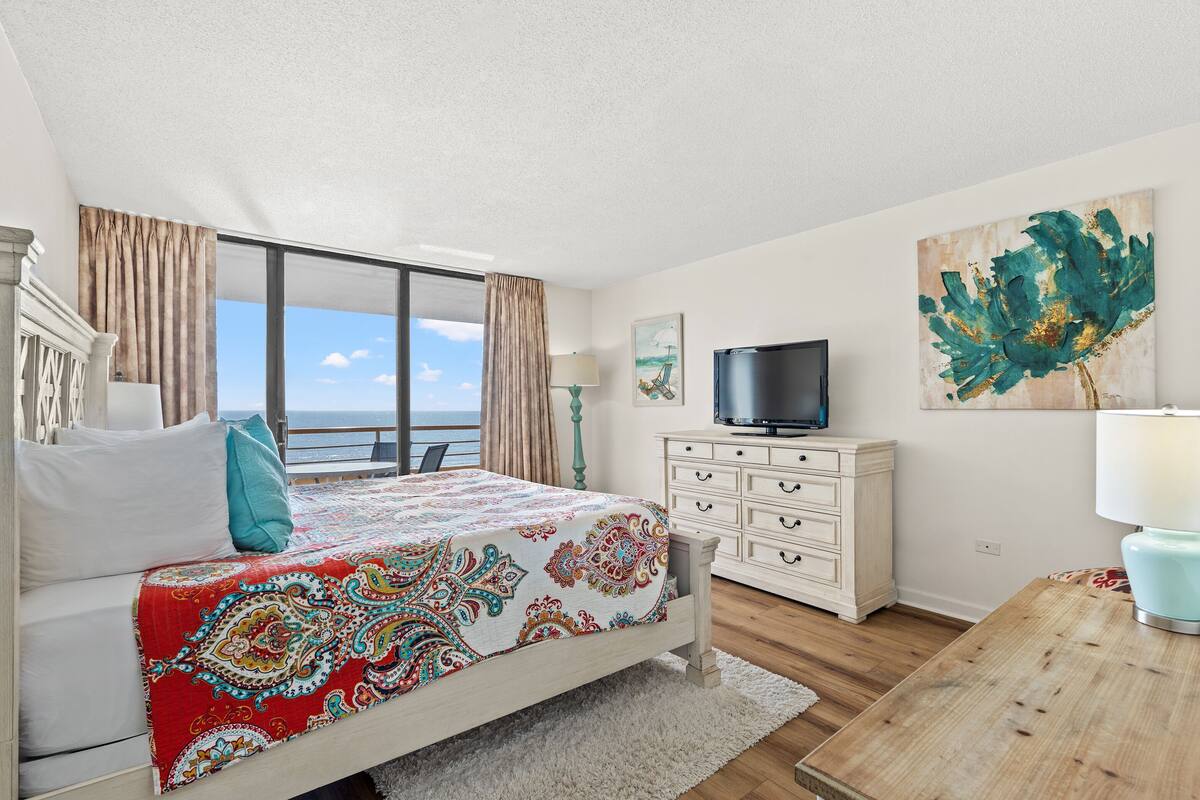 A bedroom with a bed, a TV, a dresser, a lamp, and a painting.