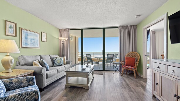 A modern living room with a sofa, coffee table, and dining area. There is a view of the beach through large windows. - Ocean Creek Resort (Myrtle Beach)