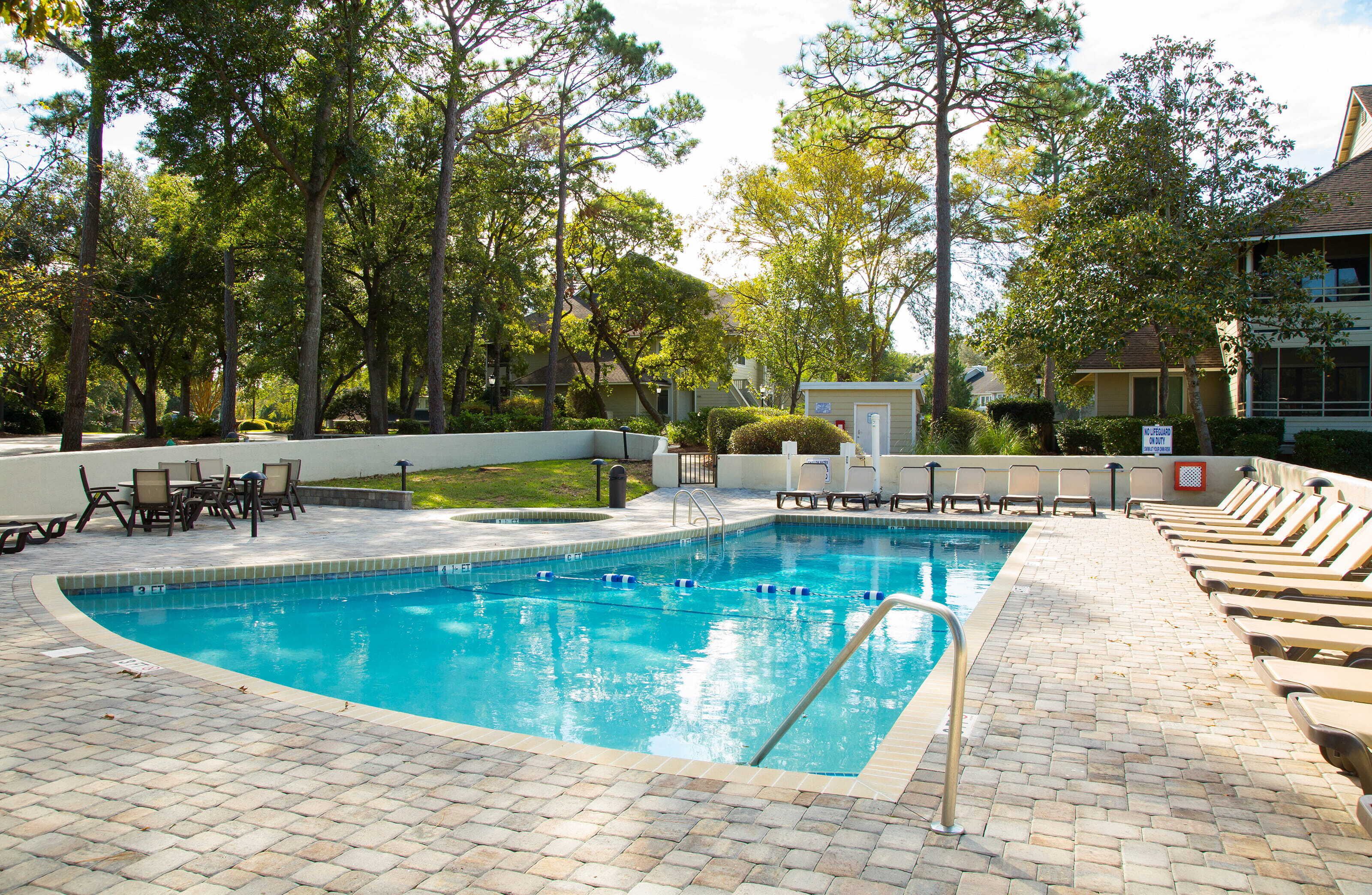 Uma piscina retangular com deck ladrilhado, cercada por árvores e um edifício.