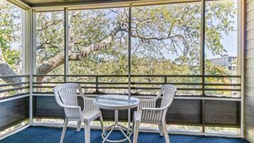 A covered patio with a table and two chairs, large windows with a view of trees and buildings.