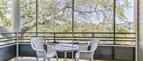 Un patio techado con una mesa y dos sillas, ventanas amplias con vista a árboles y edificios.