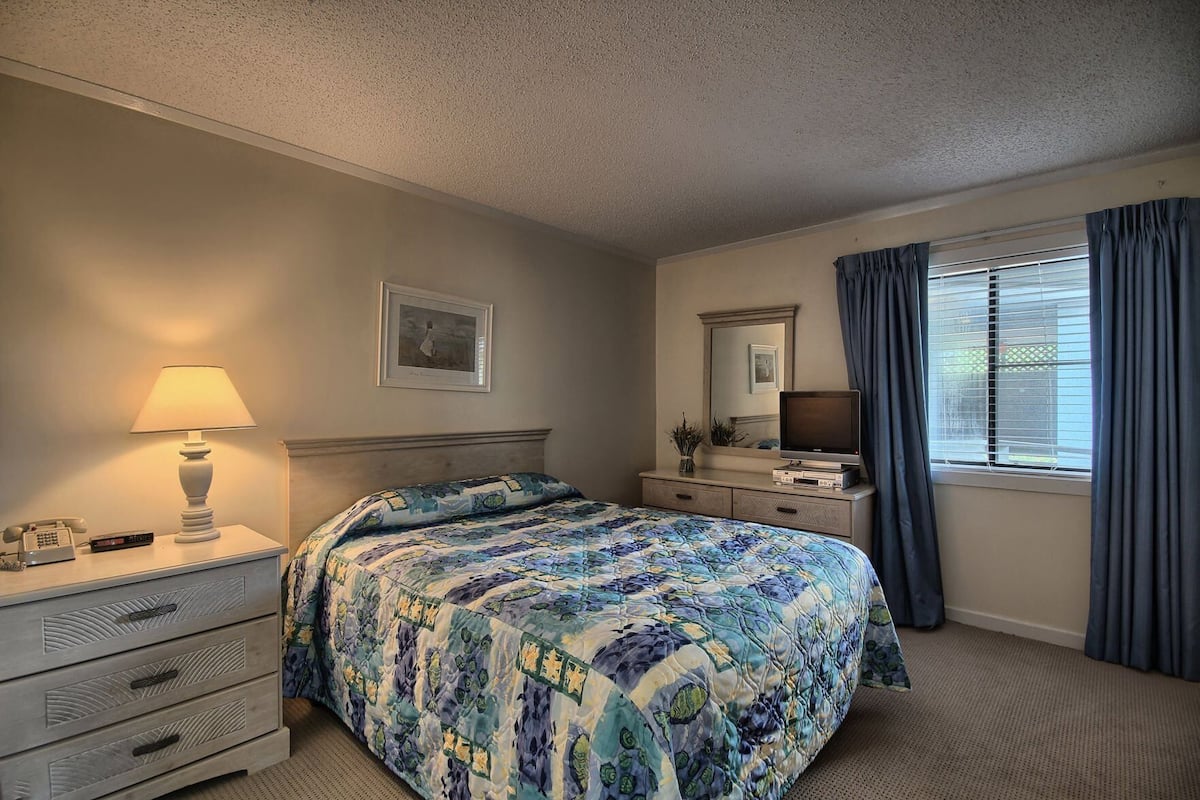A bedroom with a bed, bedside table, lamp, television, dresser, and window with curtains.